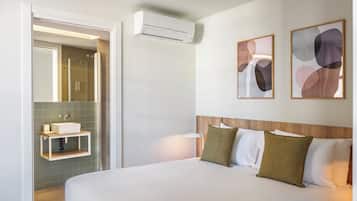 Habitación de hotel con cama, mesita de noche, aire acondicionado fijado a la pared, estante pequeño con artículos de aseo y arte abstracto en la pared.