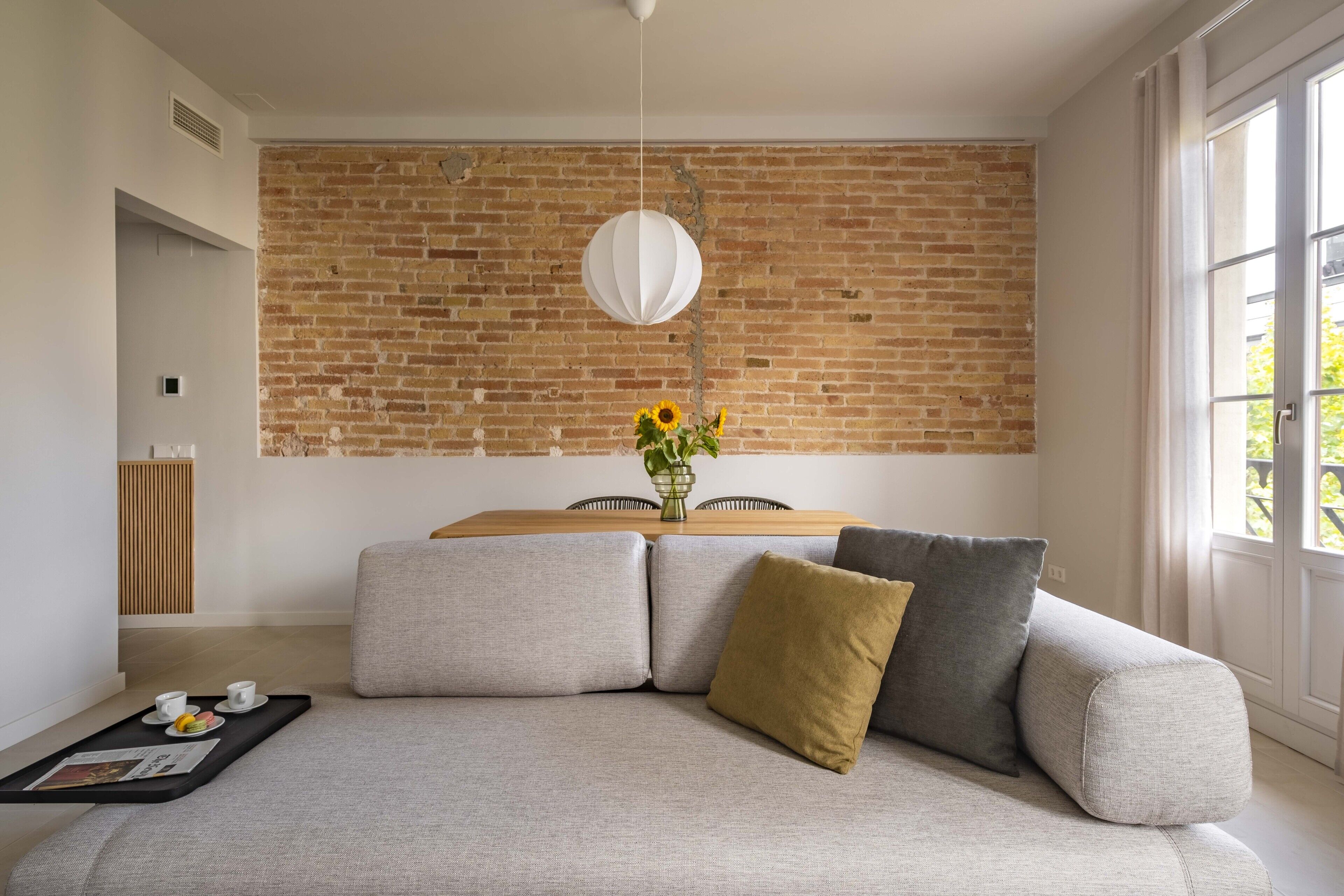 Una sala de estar moderna con pared de ladrillo, un sofá con cojines, una mesa de comedor con sillas, y una lámpara colgante.