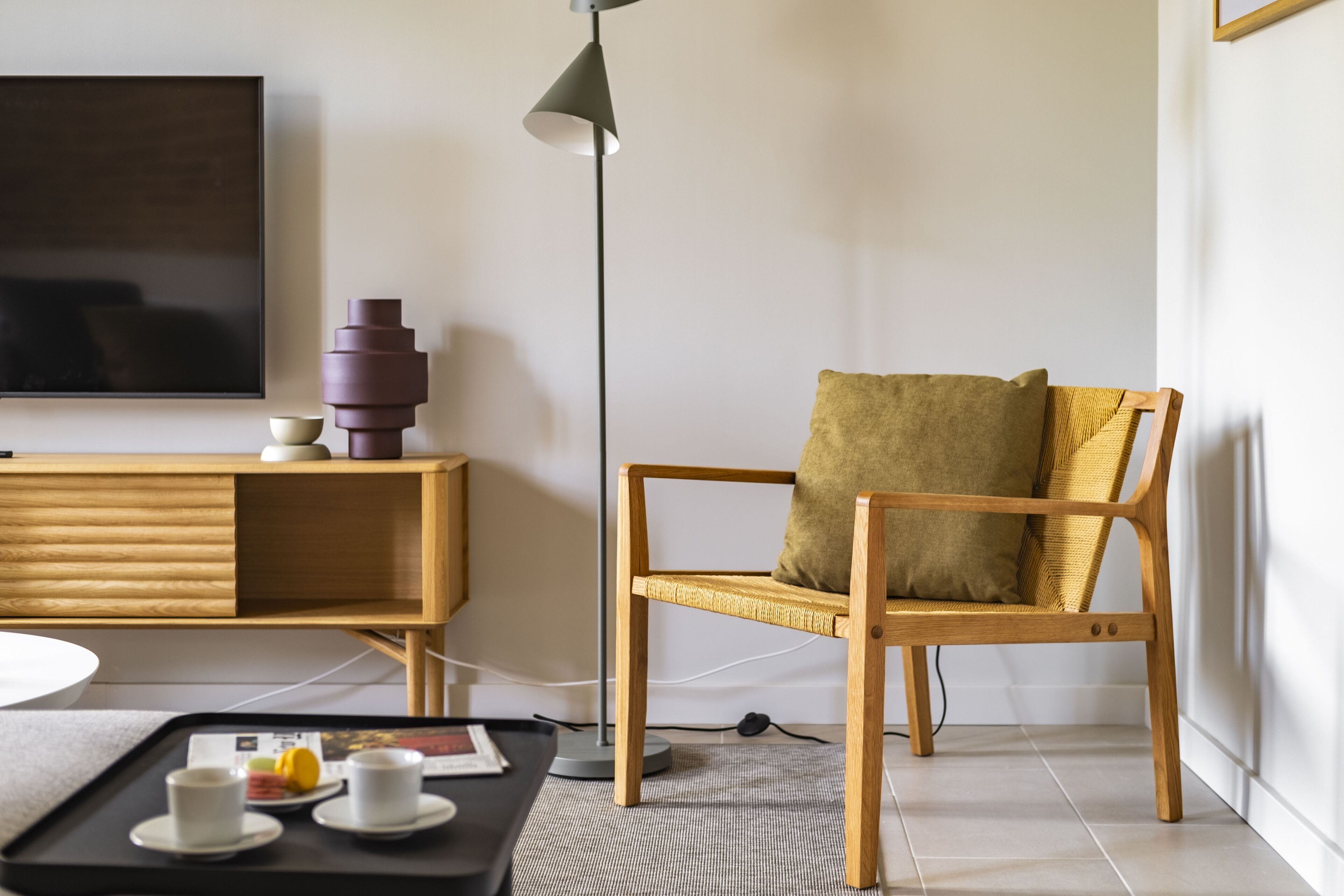 Una sala de estar moderna con una silla de madera, una bandeja negra con tazas, una lámpara de pie y un televisor de pantalla plana en la pared.