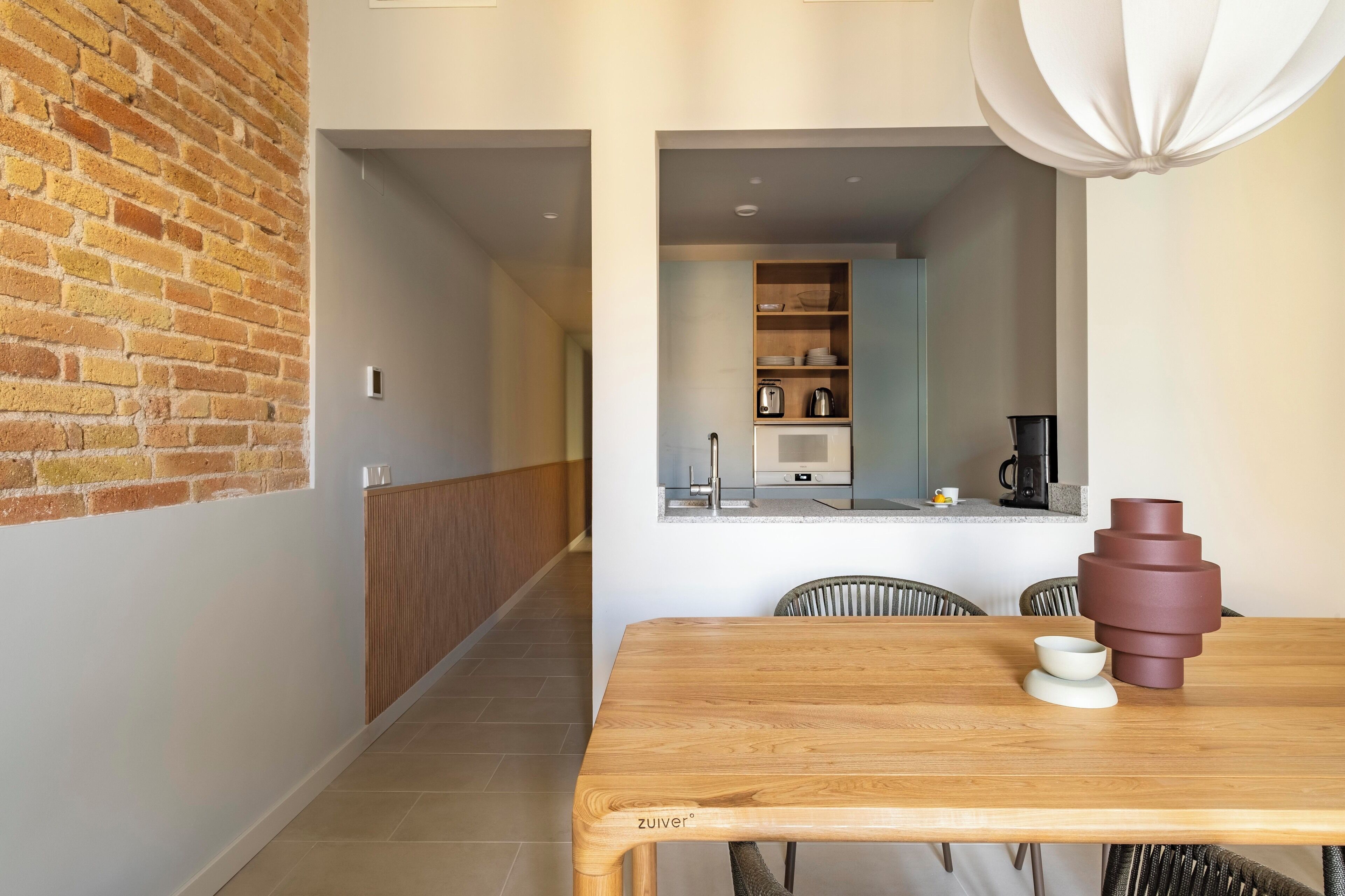 A modern kitchen with a wooden table and chairs, a brick wall, and a view into the dining area.