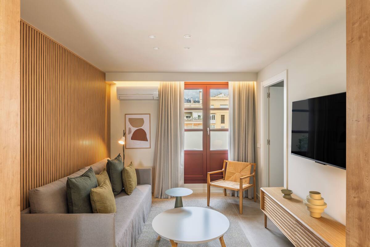 A modern living room with a sofa, a round coffee table, a wooden chair, a TV, and a window with curtains.