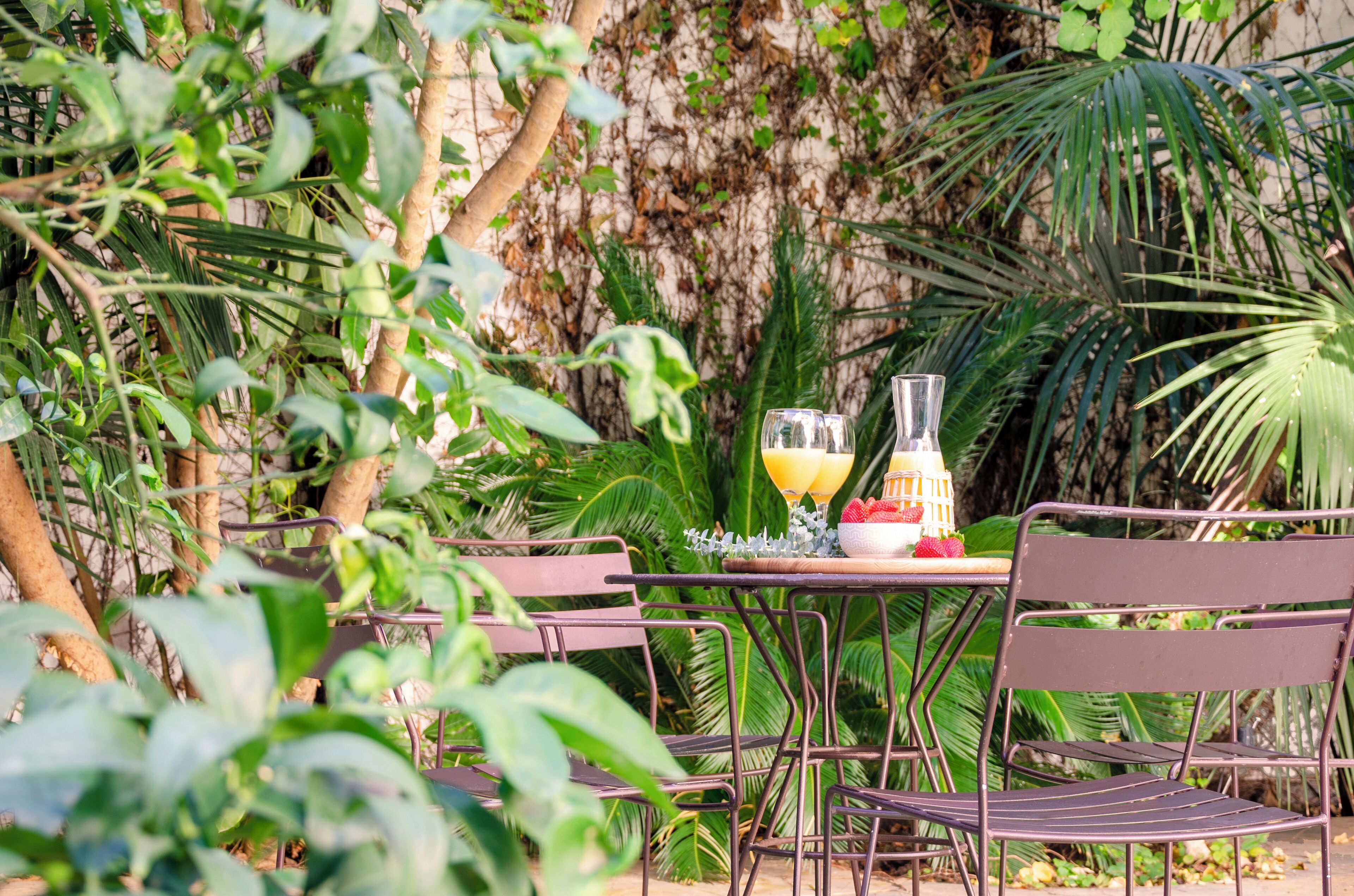 An outdoor dining area with metal chairs and a table, surrounded by lush greenery and a stone wall.
