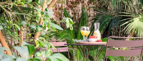 An outdoor dining area with metal chairs and a table, surrounded by lush greenery and a stone wall.