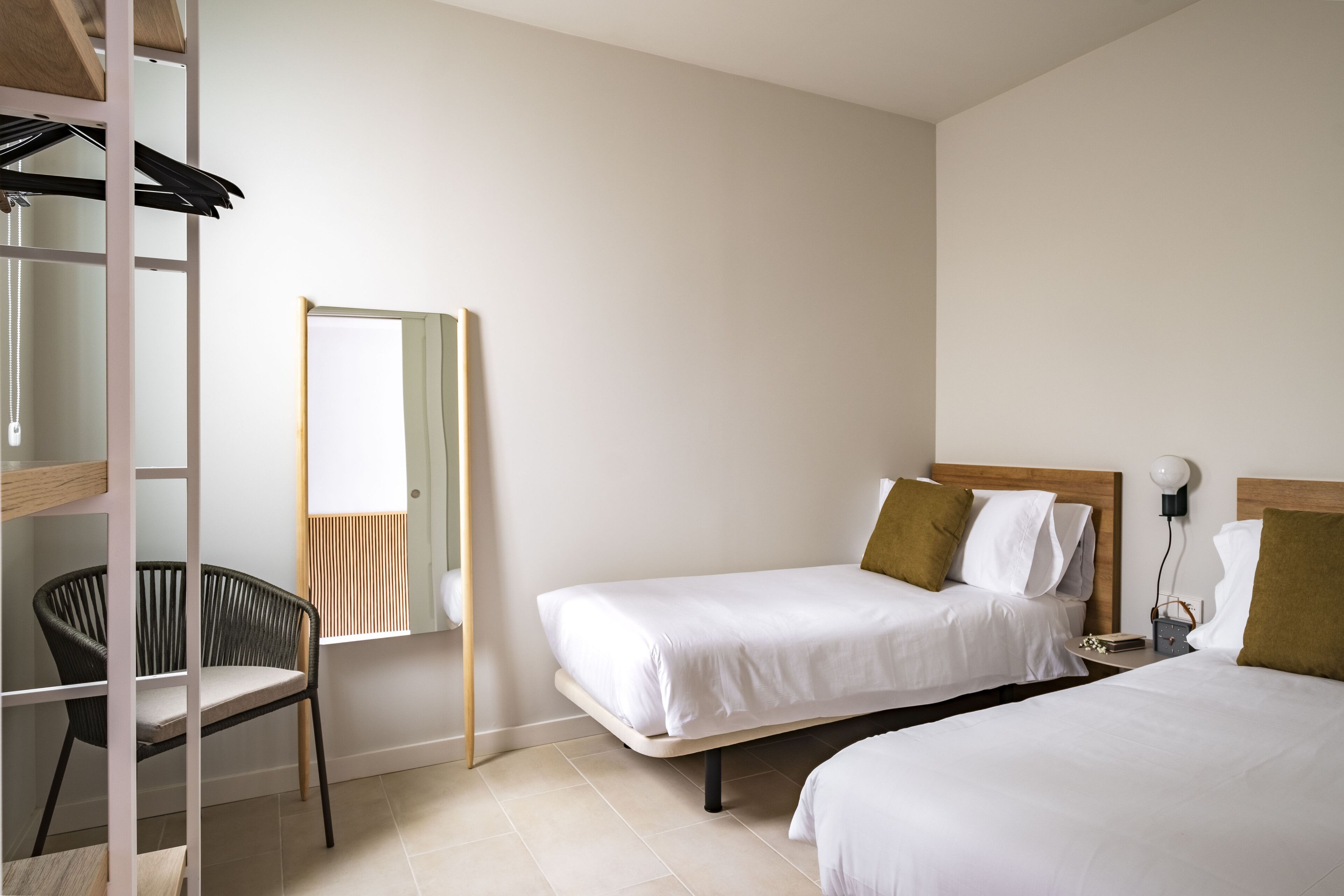 Una habitación de hotel con dos camas individuales, una silla, una mesita de noche con lámpara y un espejo en la pared.