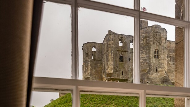 Blick auf ein Schloss durch ein Fenster, mit einem grasbewachsenen Hügel im Vordergrund.