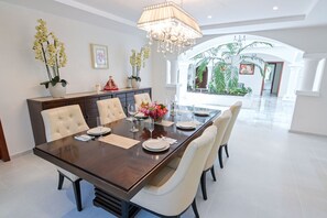 A dining area with a long table, six chairs, and a chandelier. There are potted plants and a statue on the table.