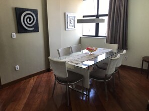 Uma sala de jantar com mesa de vidro, cadeiras de vime e uma tigela de frutas sobre a mesa.