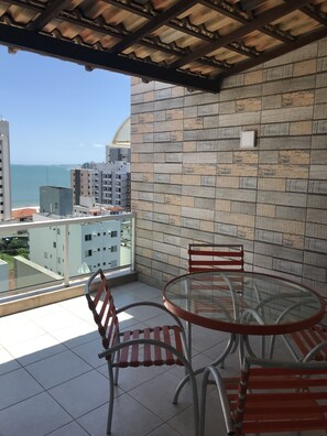 A balcony with a glass table and chairs, a tiled floor, and a view of the sea and buildings.