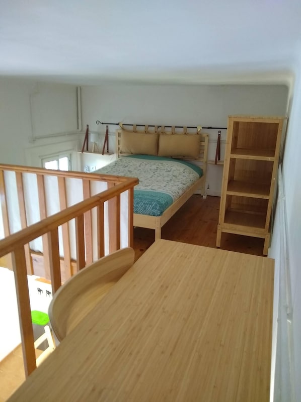 Quarto com cama, escrivaninha de madeira, cadeira, prateleira de madeira e janela.