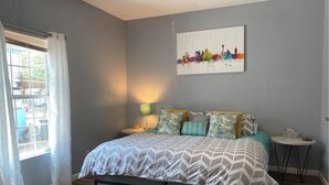 Un dormitorio con cama, mesita de noche, lámpara, una pequeña mesa y un cuadro de un paisaje urbano en la pared.