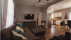 Una sala de estar con un sofá, un reposapiés y un televisor sobre un mueble bajo. Se ve una silla y un comedor al fondo.