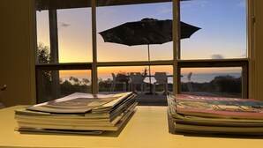 Blick durch ein Fenster auf einen Sonnenuntergang, ein Tisch mit Zeitschriften und ein Sonnenschirm.