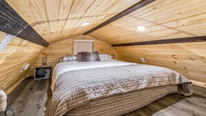 Ein gemütliches Schlafzimmer im Dachgeschoss mit Holzwänden, einem Bett mit gestreifter Decke, einem kleinen Ventilator und einem Fenster mit Jalousien.