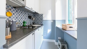 A compact kitchen with a microwave, stove, and sink. There is a window with a view of the outside, and a small wooden stool near the window.