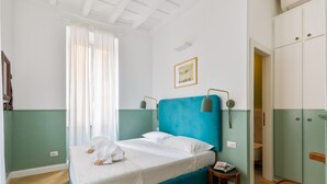 A bedroom with a teal bed, white bedding, a small bedside table with a magazine, and a wooden chair.