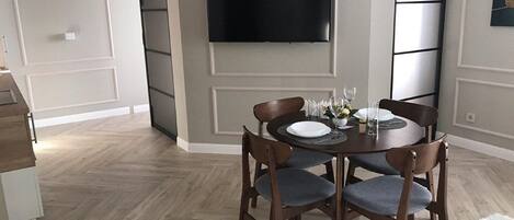 A dining area with a round table, four chairs, a flat-screen TV mounted on the wall, and a herringbone-patterned floor.