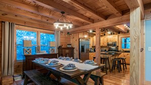 Interior de una cabaña de madera con una mesa de comedor preparada para seis comensales, una zona de cocina con electrodomésticos modernos y grandes ventanales que ofrecen vistas al exterior.