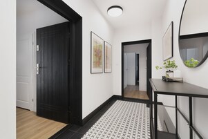 Un pasillo con una puerta negra, una pared blanca con dos cuadros enmarcados, una consola negra con un espejo y una alfombra estampada.