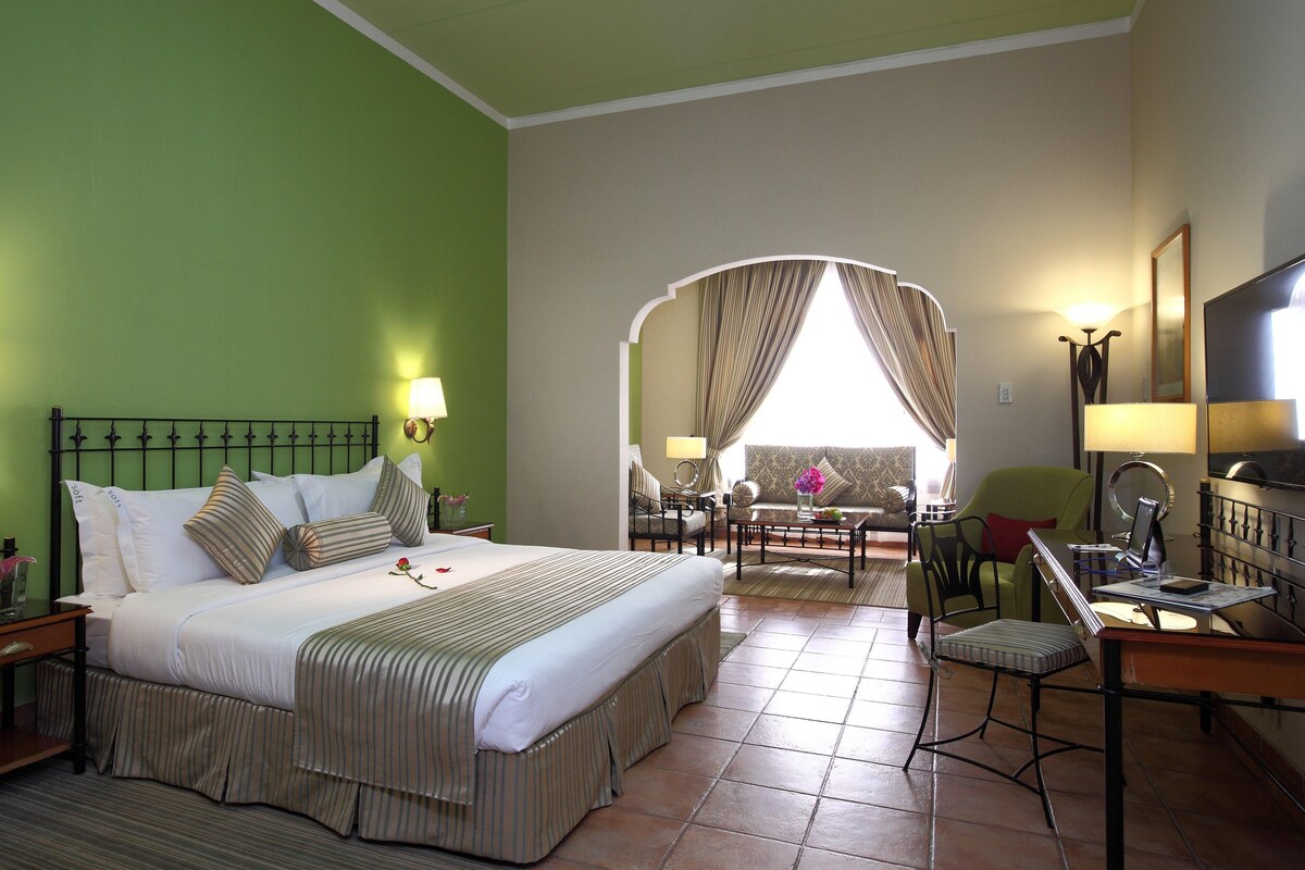 A hotel room with a large bed, a desk with a chair, a television, and a view through an archway to a balcony.