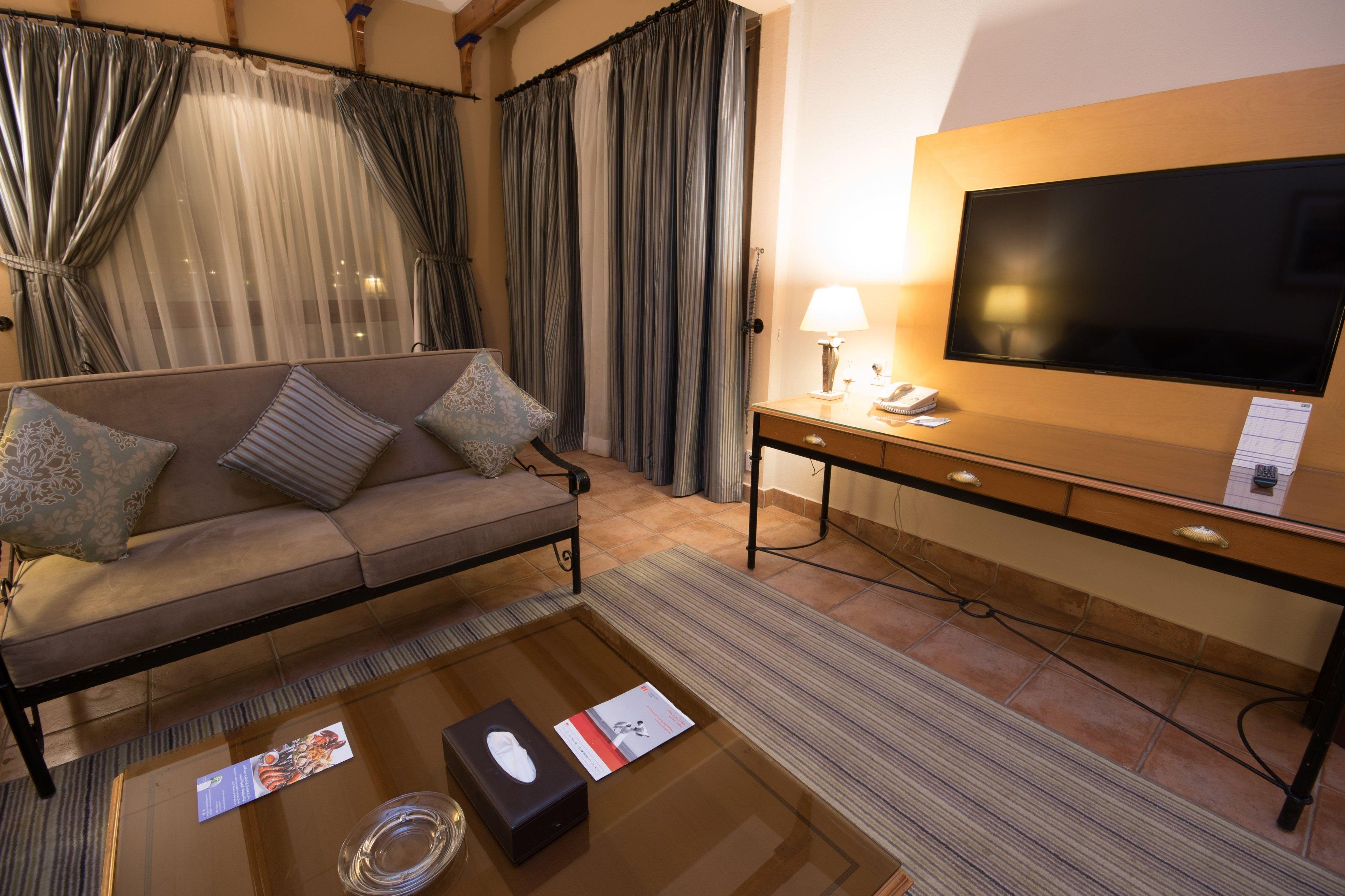 Une chambre d’hôtel équipée d’un canapé, d’une télévision, d’un bureau avec une lampe et d’une table basse avec une télécommande et une brochure.