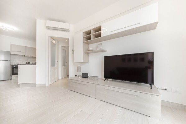 A modern living room with a large flat-screen TV, built-in shelves, and a kitchen area in the background.