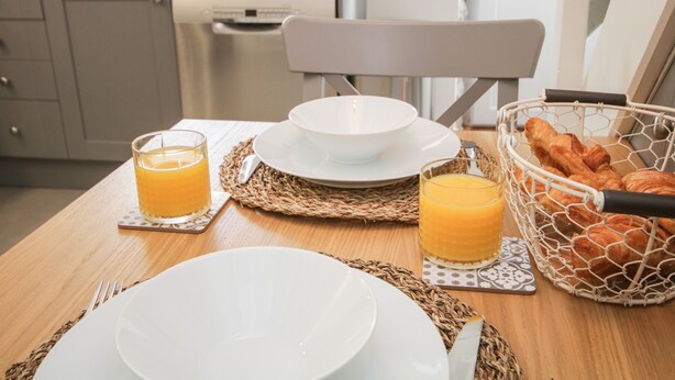 Mesa de desayuno lista con platos, vasos de jugo de naranja y una cesta de croissants.