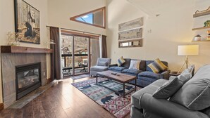 A spacious living room with a fireplace, a sofa set, a coffee table, and a large window overlooking a patio. - Enjoy Incredible Mountain Views from Private Balcony, Shared Pool & Hot Tub! (Keystone)
