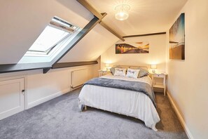 Une chambre avec une lucarne, un lit recouvert d’une couverture grise, deux tables de chevet avec des lampes et un tableau accroché au mur.