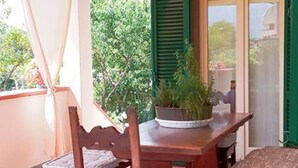 Eine überdachte Terrasse mit einem Holztisch und -stühlen, einem Blumentopf und einer Tür mit grünen Fensterläden.