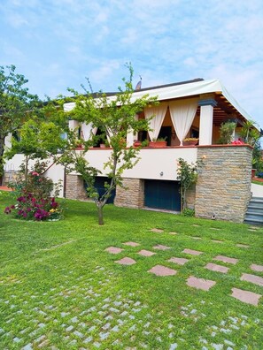 A modern two-story house with a stone facade, a well-maintained lawn, and a paved walkway leading to the entrance. - Villa El Rosal (Pietrasanta)