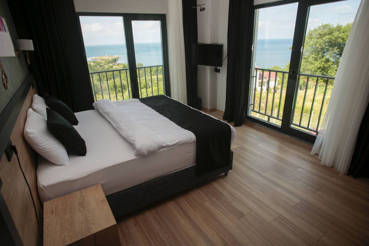Ein Hotelzimmer mit einem großen Bett, einem Holznachttisch, einem Flachbildfernseher und einem Balkon mit Meerblick.