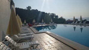 Zona de piscina con sillas de sol y sombrillas, rodeada de árboles y cielo despejado.
