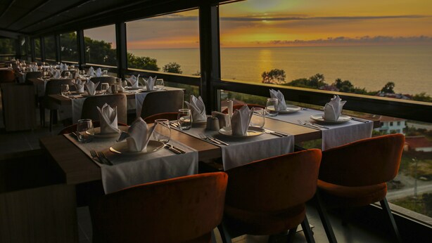 Un restaurante con mesas listas para comer, amplias ventanas que ofrecen vista del mar al atardecer.