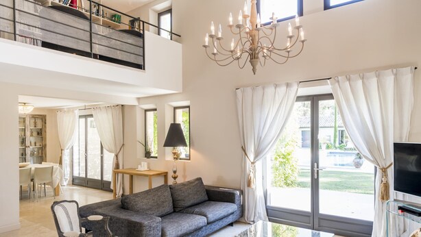 A spacious living room with a grey sofa, a glass coffee table, and a chandelier. There is a TV on a stand and a dining area in the background. - Vacation rental house with pool LOURMARIN LS2-385 (Lourmarin)