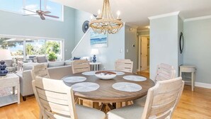 Un comedor amplio con una mesa redonda de madera rodeada por seis sillas, una araña y vistas al exterior a través de ventanas grandes.