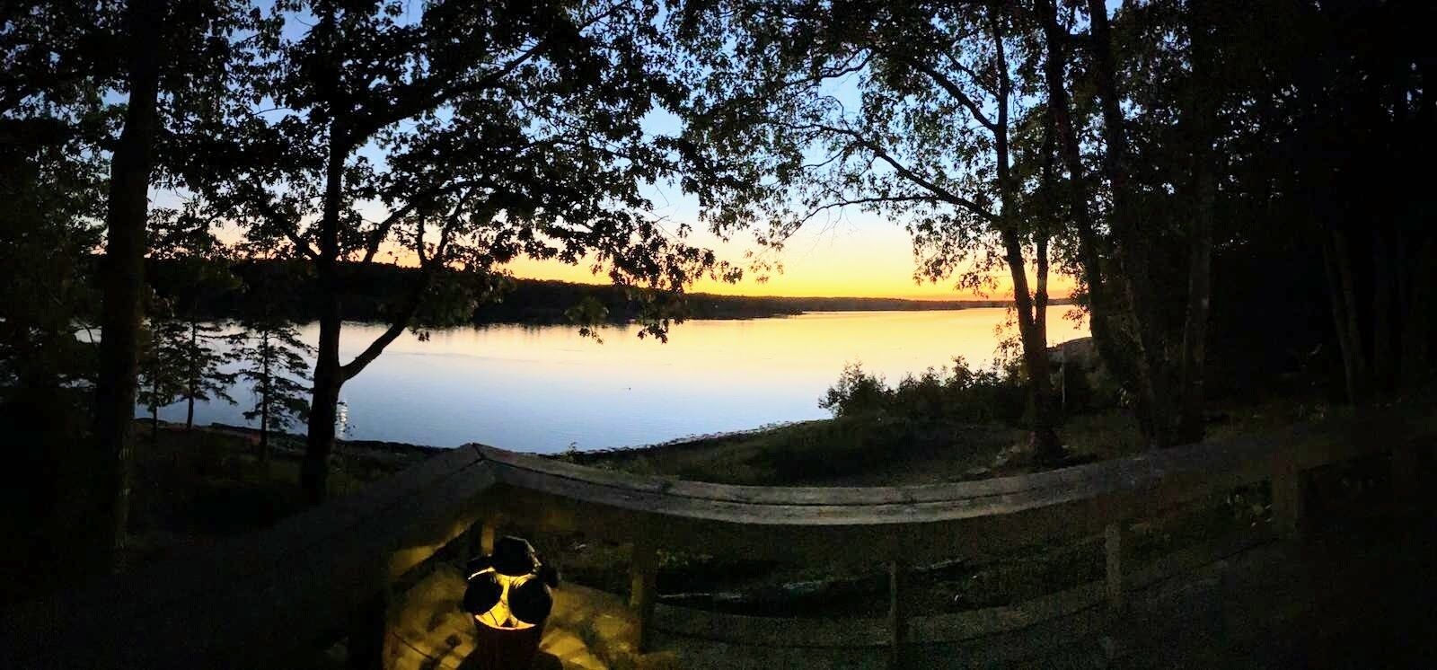 Zonsondergang uitzicht vanaf een houten terras, met uitzicht op een rustig meer, omgeven door bomen.