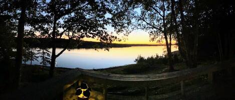 Zonsondergang uitzicht vanaf een houten terras, met uitzicht op een rustig meer, omgeven door bomen.
