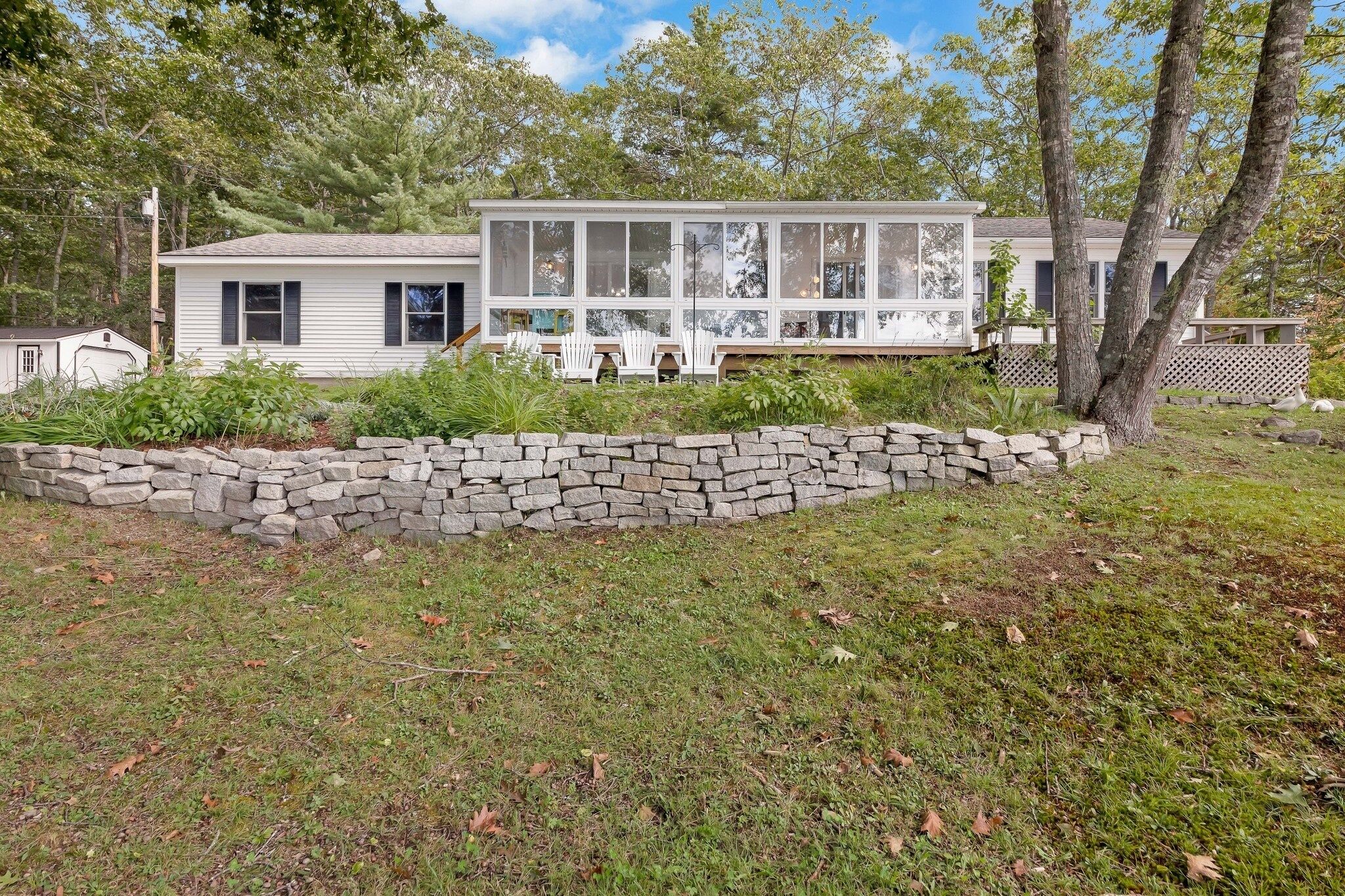 A single-story house with a large front porch, surrounded by a stone wall and lush greenery.