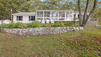 A single-story house with a large front porch, surrounded by a stone wall and lush greenery.