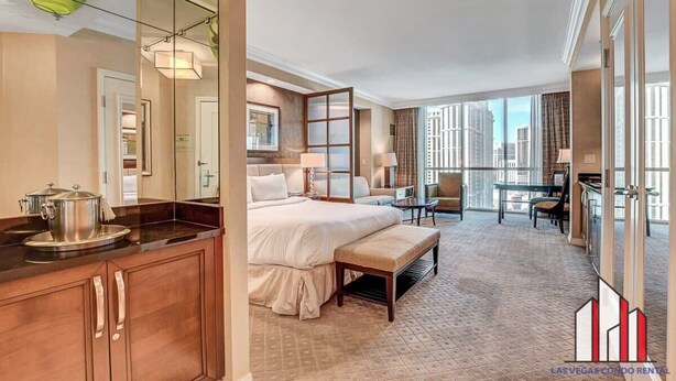 A hotel room with a large bed, a desk, a chair, a bench, a large mirror, a window with a city view, and a kitchenette with a stove.