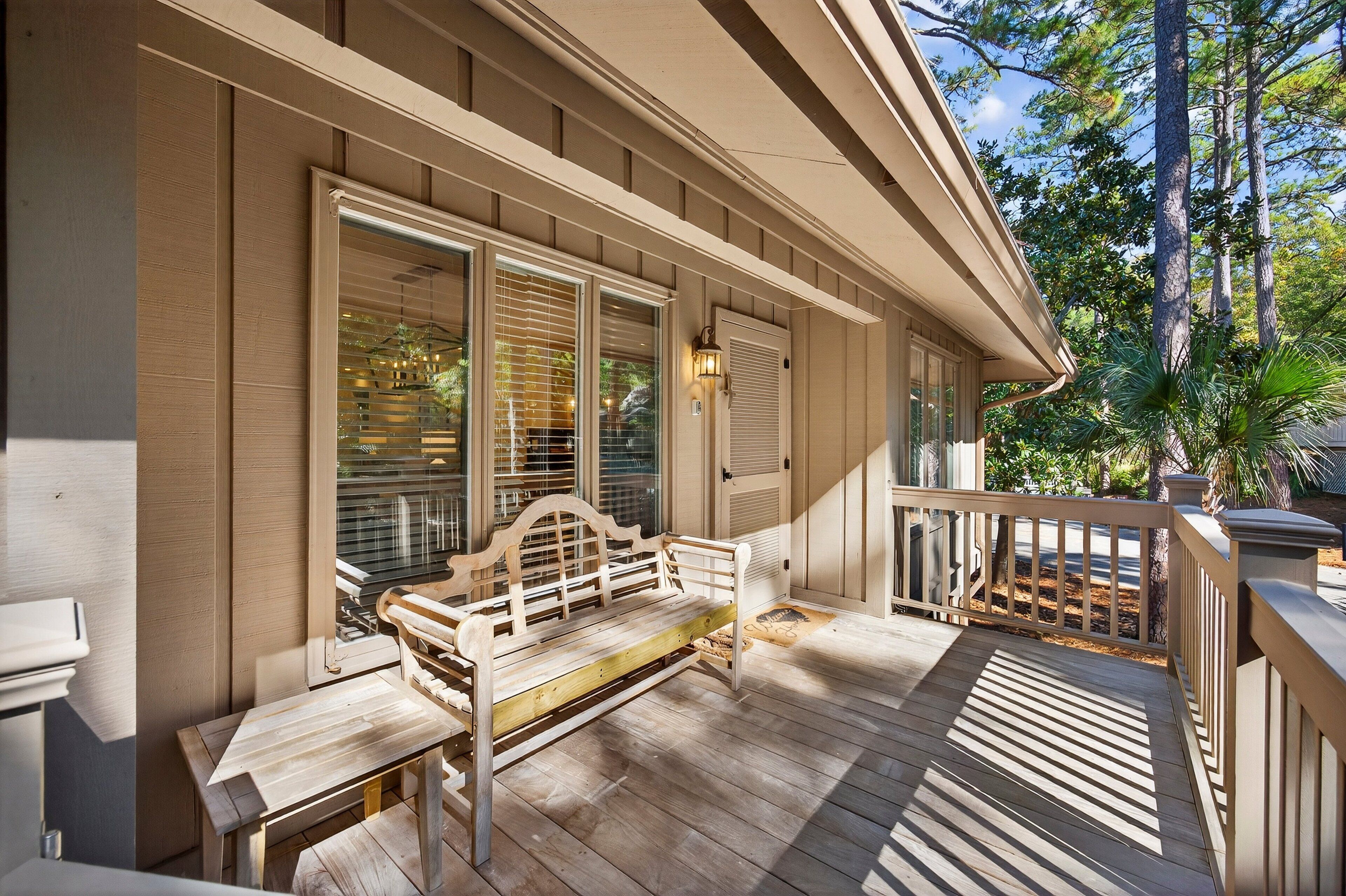 Une terrasse en bois avec un banc et une table, une véranda couverte avec une balustrade, et une vue sur les arbres et le ciel.