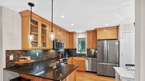 A modern kitchen with wooden cabinets, a granite countertop, stainless steel appliances, and a dining area with a white table and two stools. - River Views at Inlet Cove! Community Dock and Pool (Seabrook Island)