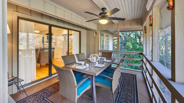A covered outdoor dining area with a table set for four, wicker chairs with blue cushions, a ceiling fan, and a view of greenery through large windows. - River Views at Inlet Cove! Community Dock and Pool (Seabrook Island)