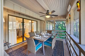 A covered outdoor dining area with a table set for four, wicker chairs with blue cushions, a ceiling fan, and a view of greenery through large windows. - River Views at Inlet Cove! Community Dock and Pool (Seabrook Island)