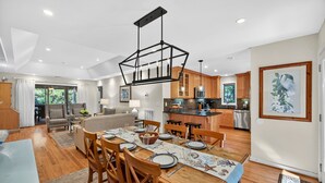 A spacious living area with a wooden dining table set for a meal, a kitchen with modern appliances, and a large window offering a view of greenery. - River Views at Inlet Cove! Community Dock and Pool (Seabrook Island)