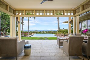 A covered outdoor area with wicker furniture, a table set for dining, and a view of a swimming pool and ocean. - Exquisite 4BR Hainoa Estate 122 at Hualalai – Ocean Views & Infinity-Edge Pool (Kailua Kona)