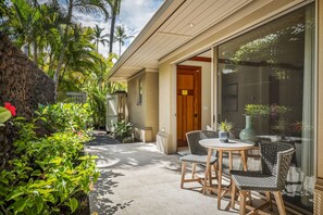 A patio with wicker chairs and a table, surrounded by lush greenery and palm trees. - Exquisite 4BR Hainoa Estate 122 at Hualalai – Ocean Views & Infinity-Edge Pool (Kailua Kona)