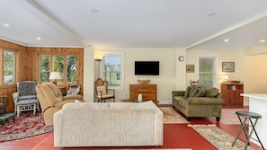 A spacious living room with a large sofa, two armchairs, a coffee table, and a television. There are wooden paneled walls, a red carpet, and a window with blinds.