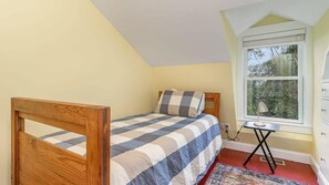 A bedroom with a wooden bed, a checkered bedspread, a small table with a lamp, and a window with a view of trees.