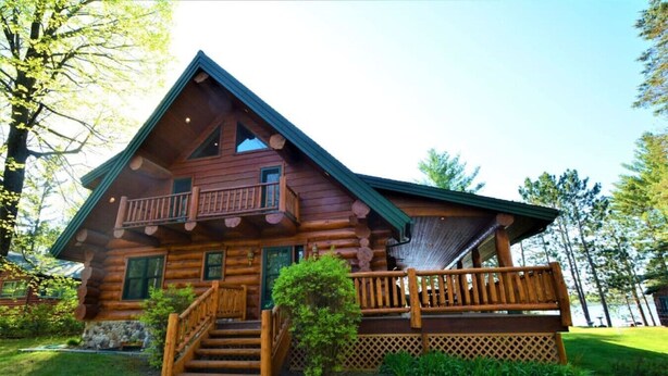 A log cabin with a balcony, a staircase leading to the entrance, and a surrounding grassy area. - Large Home- Water Toys, Dock, Deck, Game Room (Brainerd)